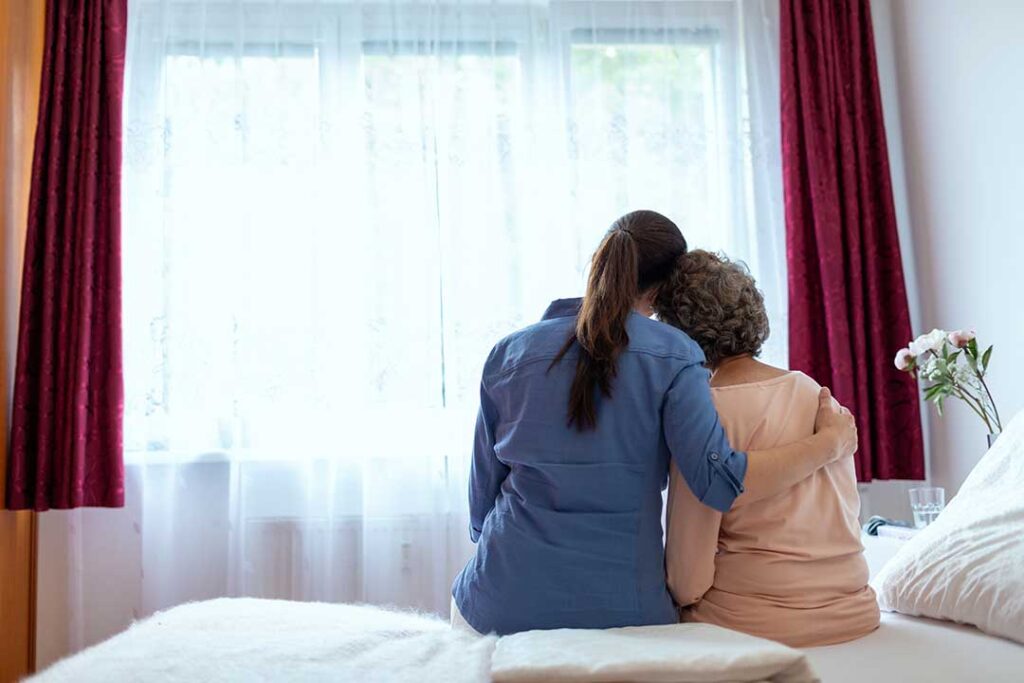 A middle-aged adult and elderly parent sit together holding hands, engaged in a warm, thoughtful conversation at home.