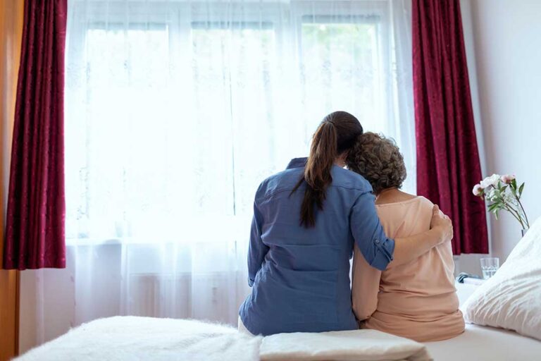 A middle-aged adult and elderly parent sit together holding hands, engaged in a warm, thoughtful conversation at home.