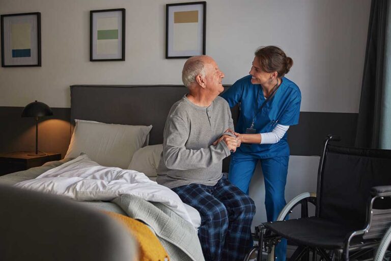 Two seniors sitting at a table with a caregiver, one in casual clothes and one in a wheelchair.